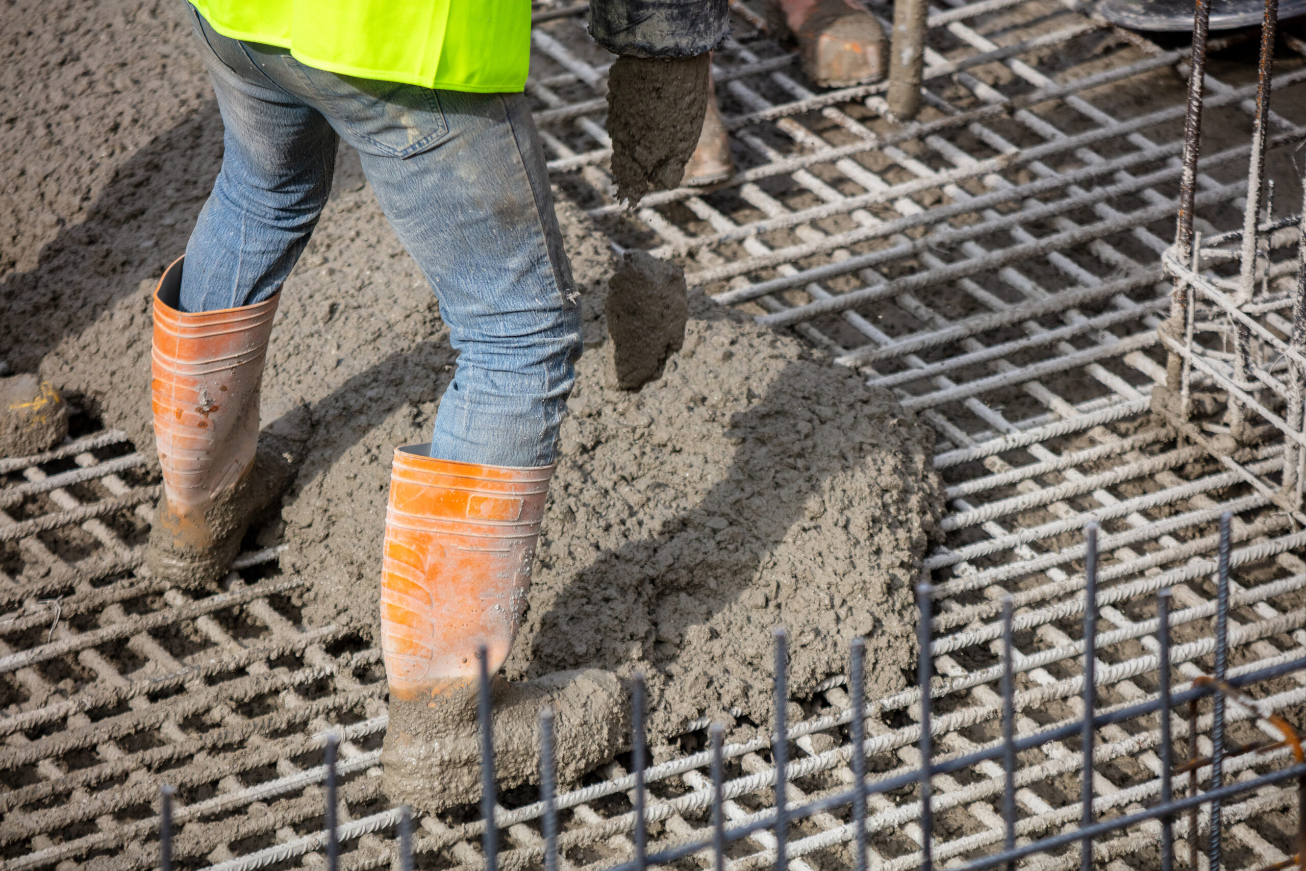 worker within orange boots pouring wet concrete over reinforced steel grid at construction site under daylight